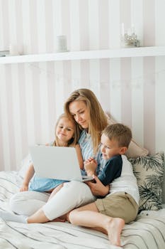 A mother sits on a bed with her children, sharing a joyful moment using a laptop.